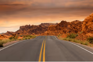 Open road leading through a desert landscape with red rock mountains at sunset.